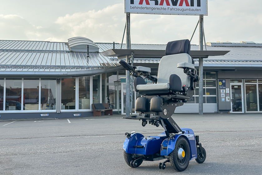 A blue electric wheelchair, the PR50, is parked in the courtyard of Paravan GmbH and has been raised slightly.