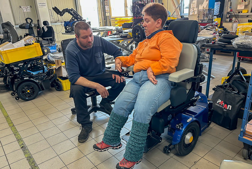 A woman in an orange sweater is sitting in a blue PR50. Next to her sits a workshop employee from Paravan’s wheelchair division, watching her. 