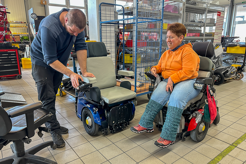 A Paravan GmbH workshop employee is working on a new blue PR50 electric wheelchair. To his right, a woman wearing an orange sweater, also in a wheelchair, is watching. 