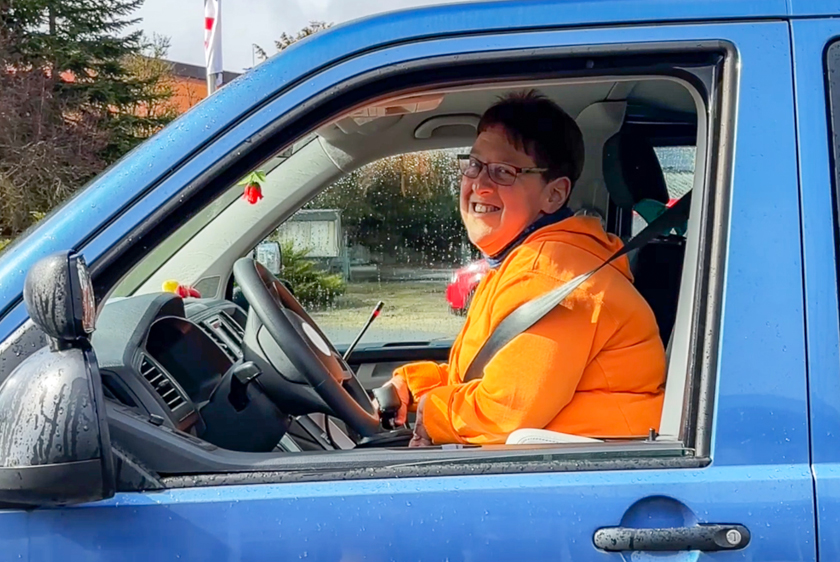 A woman in an orange jacket is smiling as she looks out the driver's side window of a blue car.
