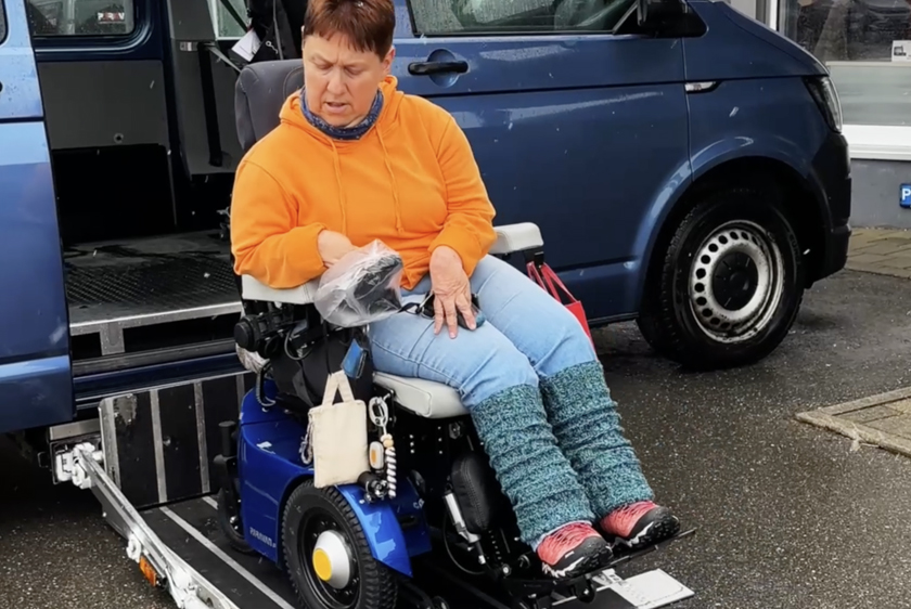 A woman wearing an orange sweater is sitting in her blue PR50 wheelchair. The wheelchair is on a cassette lift that has been extended, on the ground in front of a blue car. 