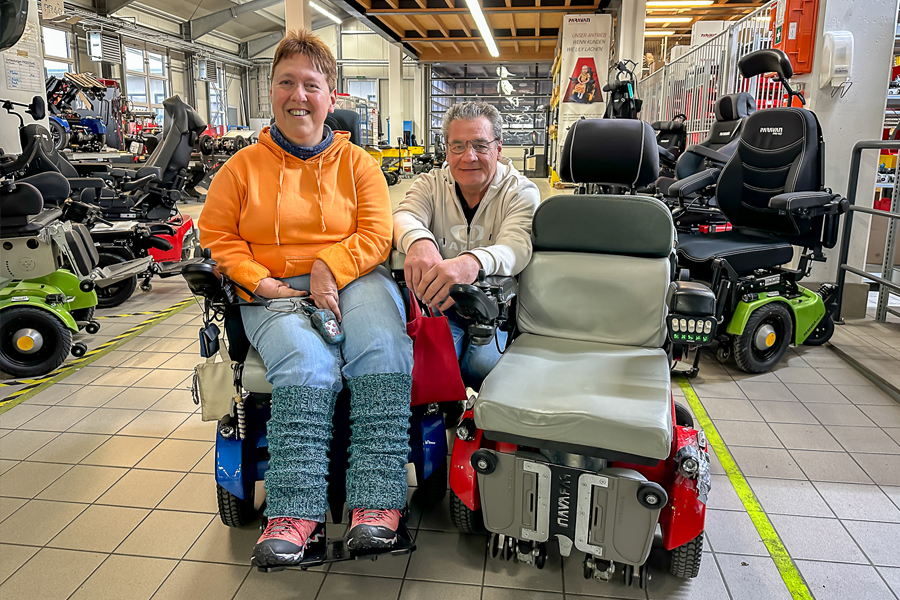 A woman wearing an orange sweater and sitting in a new blue wheelchair, with an older red wheelchair to her right, is standing in the wheelchair workshop at Paravan GmbH. The head of the wheelchair department is kneeling in the middle, behind the two wheelchairs. 
