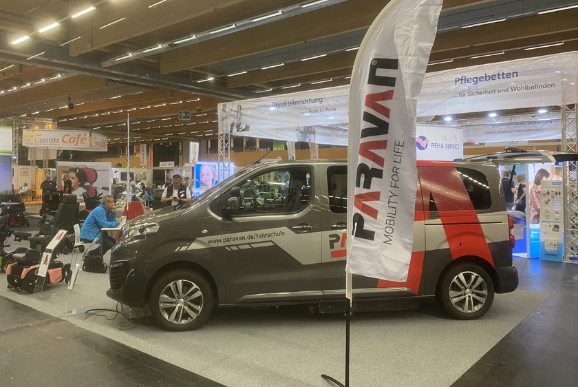 A Paravan GmbH exhibition booth with a Paravan beach flag in the center foreground. Behind it is a car, and to the left of the car are a few wheelchairs. 