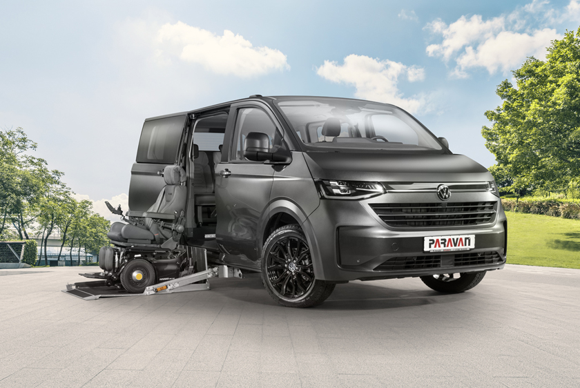 A VW Caravelle is parked in an animated setting featuring a large square and a park to its right. A black wheelchair is positioned on the extended cassette lift, with the VW Caravelle's side door open. 