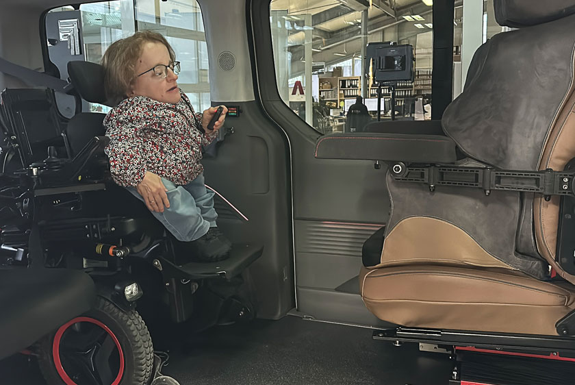 A woman with dwarfism is in the car with her wheelchair; the driver's seat is across from her, but it has been turned around so that she can transfer from her wheelchair to the driver's seat.