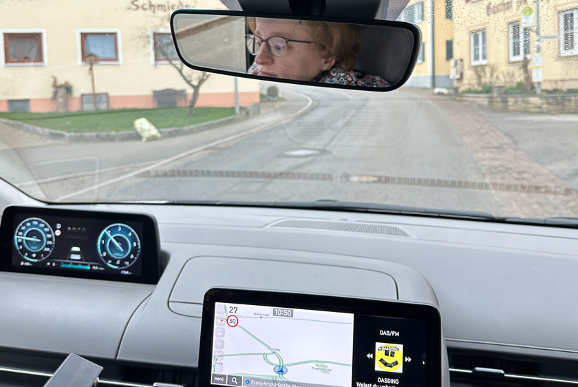 A view from inside the car, looking out the windshield at the street in a small town. In the reflection of the rearview mirror, you can see a woman deep in thought.