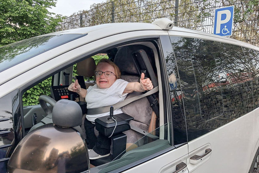 A smiling woman extends her right arm out the open driver's window, giving a thumbs-up. Her vehicle is equipped with two joysticks, a Paravan touchscreen display, and a custom-fitted driver's seat. In the upper right corner of the image, you can see a parking sign for people with disabilities.