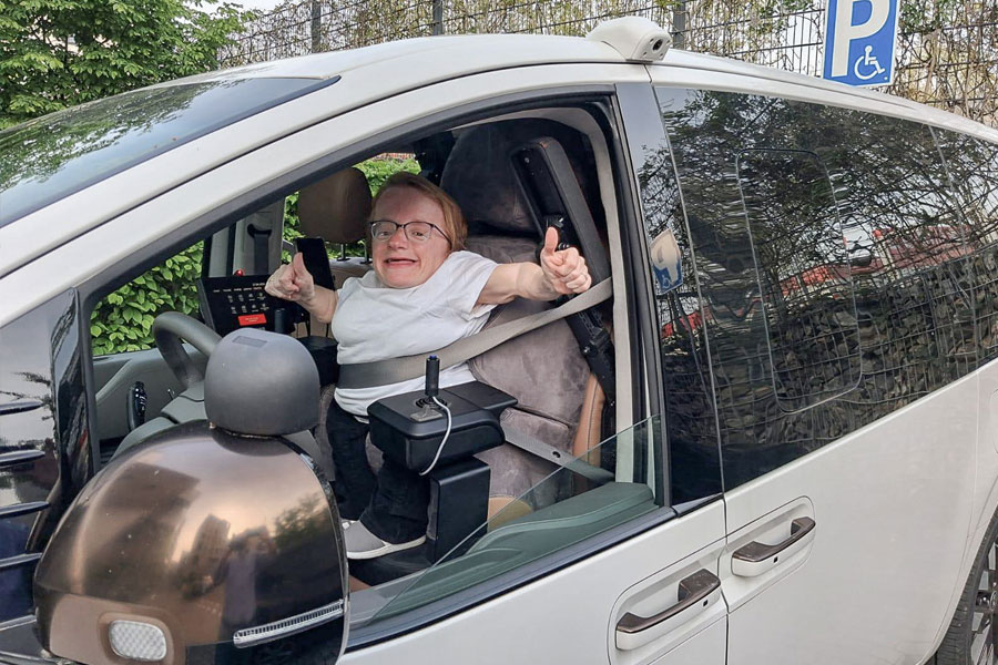 A smiling woman extends her right arm out the open driver's window, giving a thumbs-up. Her vehicle is equipped with two joysticks, a Paravan touchscreen display, and a custom-fitted driver's seat. In the upper right corner of the image, you can see a parking sign for people with disabilities.