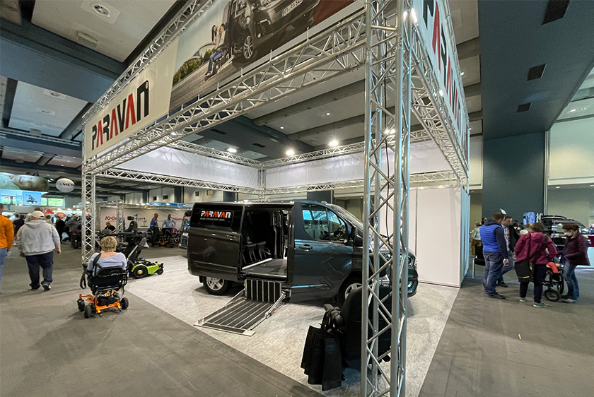 An exhibition booth with a truss system, featuring a car with the Paravan logo and wheelchairs.