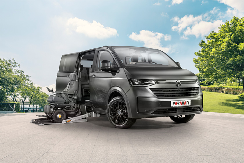 A VW Caravelle is parked in an animated setting featuring a large square and a park to its right. A black wheelchair is positioned on the extended cassette lift, with the VW Caravelle’s side door open.