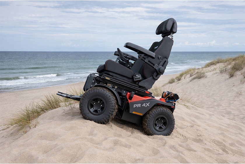 The PR4X off-road electric wheelchair is standing in the middle of the sand on a dune. The ocean is to the left. 
