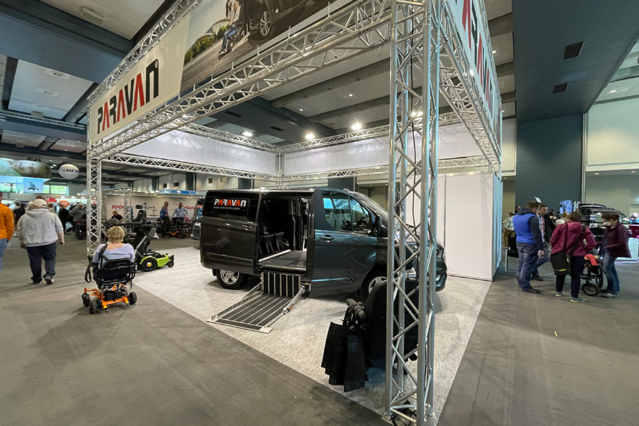 An exhibition booth with a truss system, featuring a car with the Paravan logo and wheelchairs.