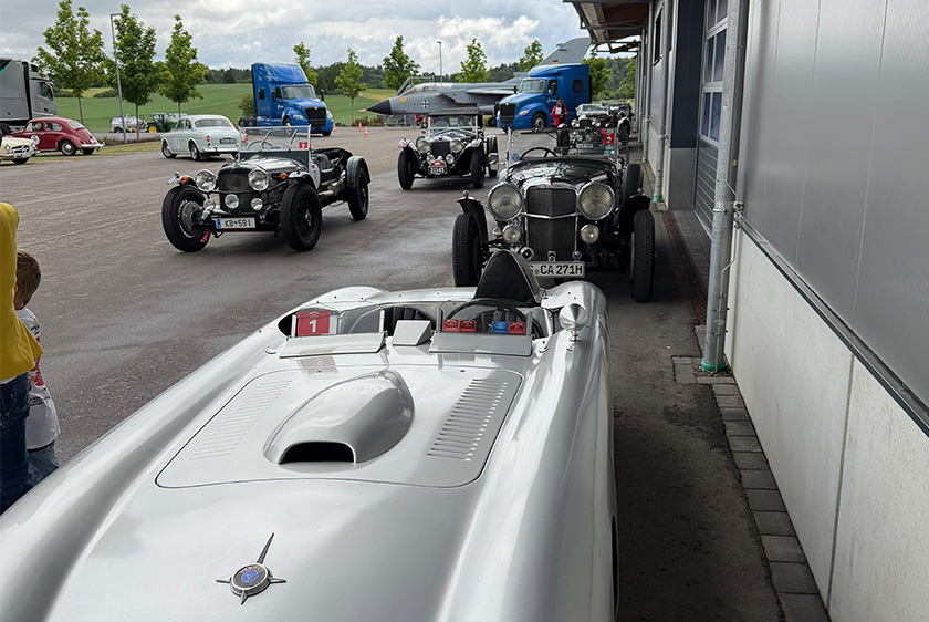 Several classic cars are parked side by side and in a row to the left of a building. In the background are two blue trucks and a Tornado.