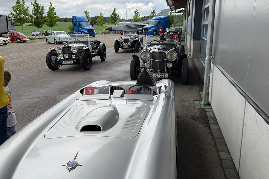Several classic cars are parked side by side and in a row to the left of a building. In the background are two blue trucks and a Tornado.