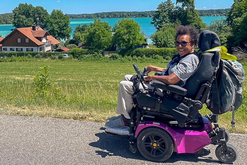 An elderly woman with a beaming smile in a pink PR30/II electric wheelchair, out and about at Lake Tegernsee amid bright sunshine and green meadows.