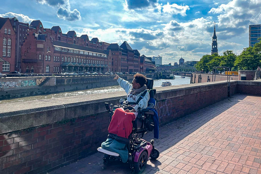 A woman in an electric wheelchair is standing by a river in a city center and pointing at a building on the other side of the river.