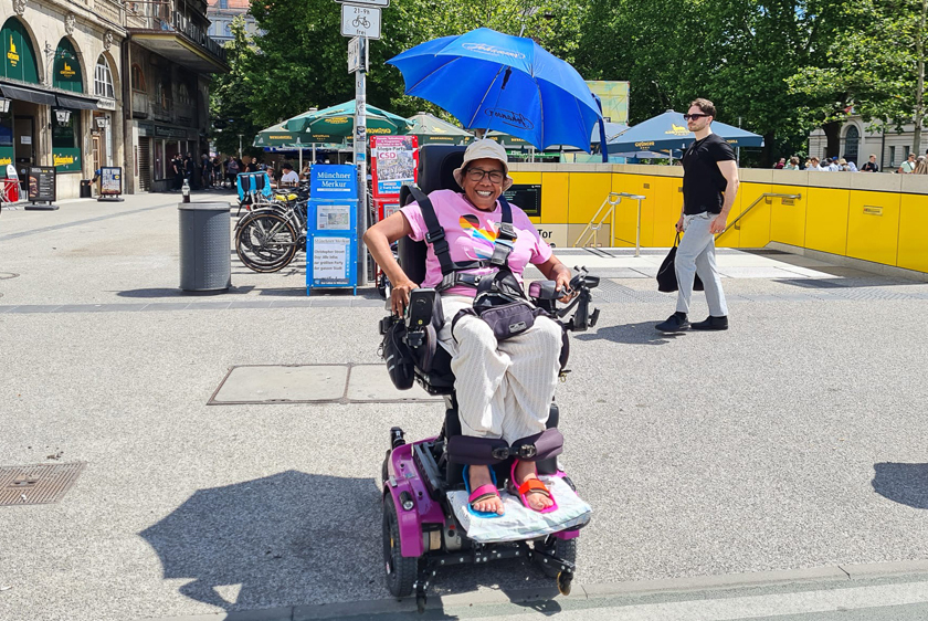 A woman with a beaming smile is sitting in her pink PR30/II electric wheelchair, which is slightly raised, in a city. An open parasol has been attached to her wheelchair. 