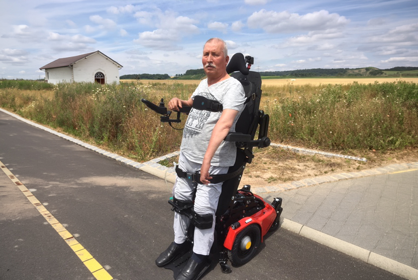 An elderly man is standing in his PR40 standing wheelchair on a path in the countryside. All around him, bathed in sunlight, are meadows and fields, and to his left is a small cottage.