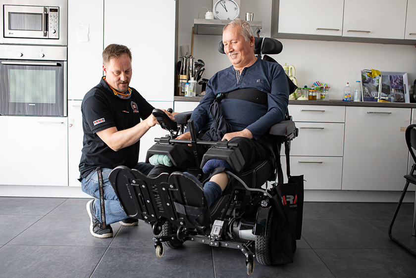 A Paravan employee is kneeling next to an elderly man sitting in an electric wheelchair. The man is beaming as our employee raises the footrest of the PR40.