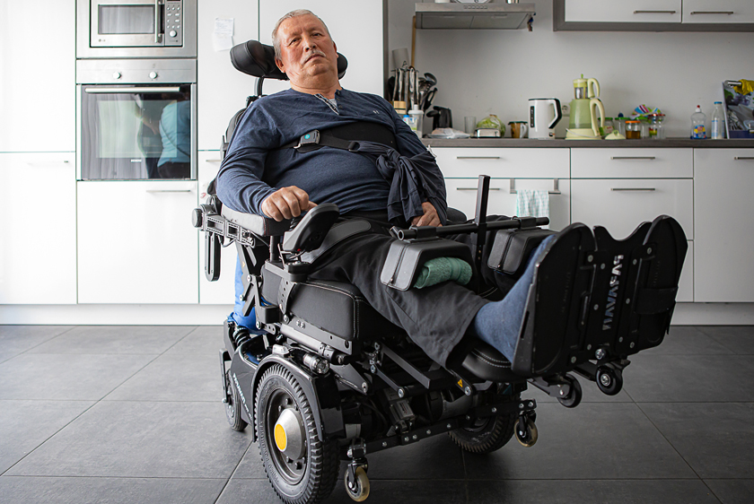 An elderly man is sitting in his PR40 electric wheelchair with his feet stretched out—that is, with the footrest raised—relaxing in his kitchen.