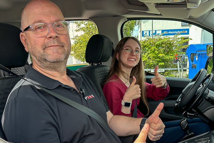 A young woman is sitting behind the wheel of a car, smiling broadly as she gives a thumbs-up. In the passenger seat sits a driving instructor from the Paravan Driving School, who is also smiling and giving a thumbs-up.