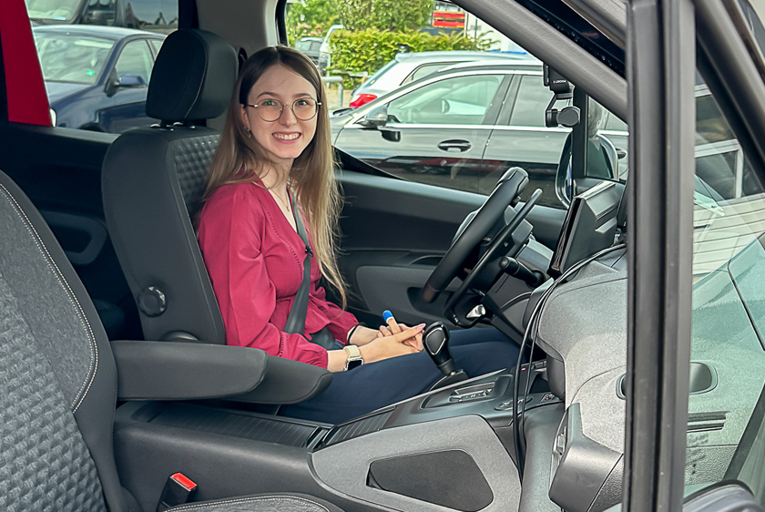 Through the open window on the passenger side of a car, a young woman wearing a wine-red top beams as she sits in the driver's seat.