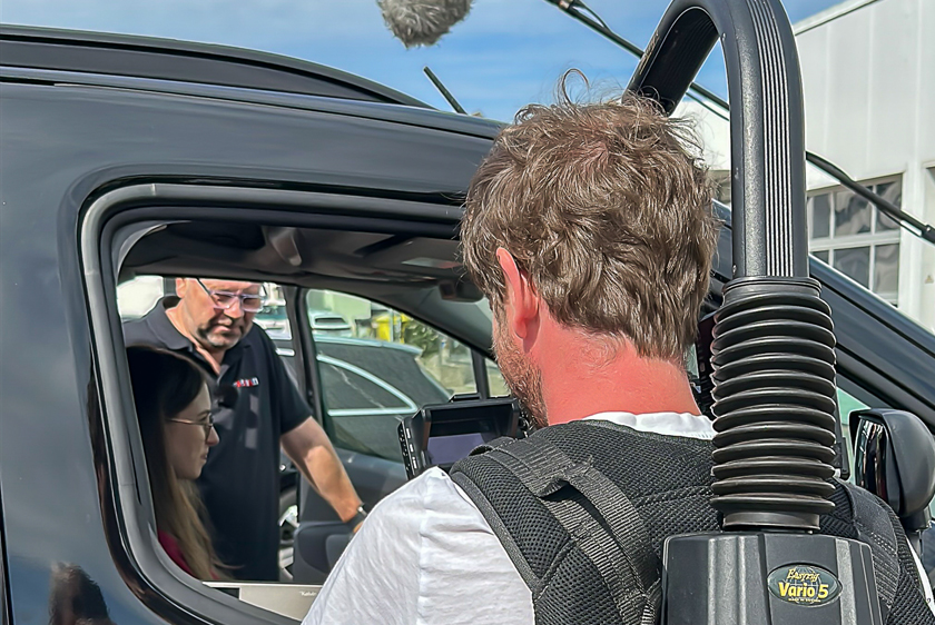 A cameraman is filming through the open passenger-side window of a car. He is filming a young woman sitting in the driver's seat; a man is standing next to her by the door, showing her something.